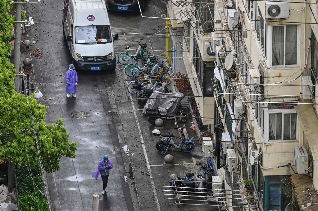 Pracovníci s osobními ochrannými prostředky (PPE) chodí po ulici vedle bytového domu během uzamčení koronaviru Covid-19 v šanghajské čtvrti Jing'an dne 23. dubna 2022.